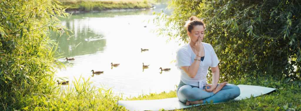 A woman sitting cross-legged on a mat beside a river, with ducks passing