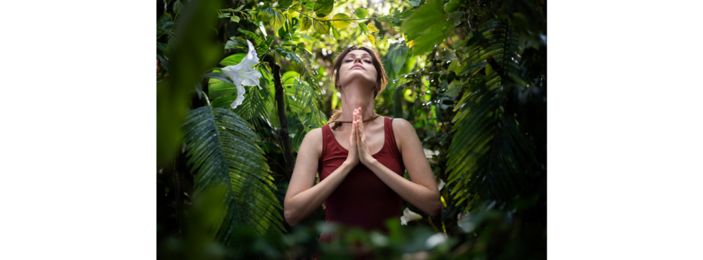 A woman standing among tropical foliage with her hands together and head tilted back