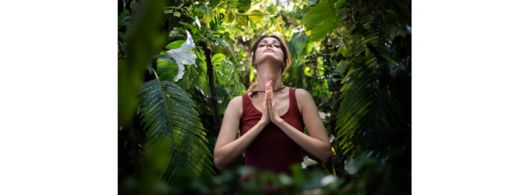 A woman standing among tropical foliage with her hands together and head tilted back