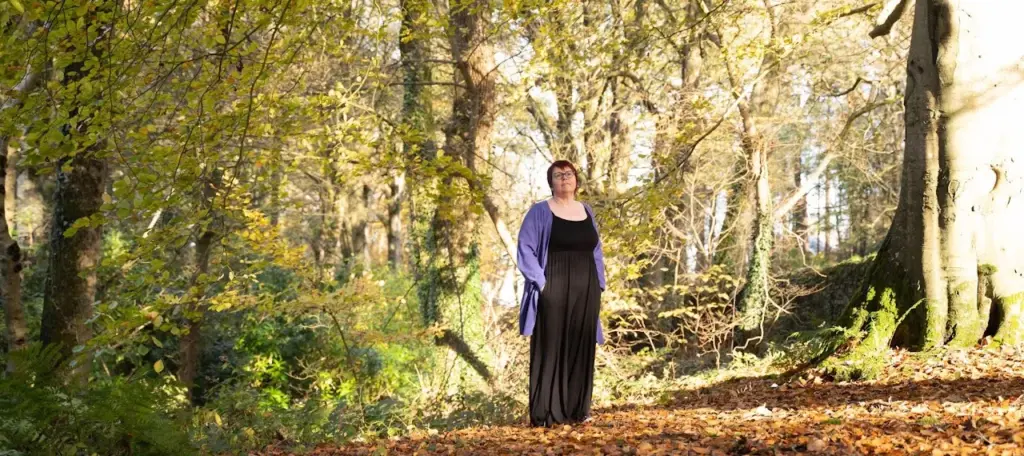 Marie Doherty standing among autumn trees in a black dress and lilac wrap