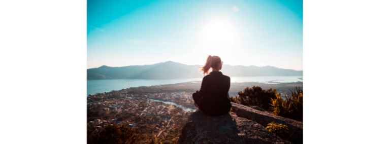 A person sitting on a hillside at sunrise, looking out over a town and the sea