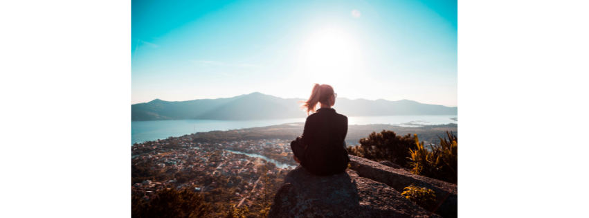 A person sitting on a hillside at sunrise, looking out over a town and the sea