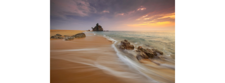 Waves washing over sand at sunset, with a rock stack offshore