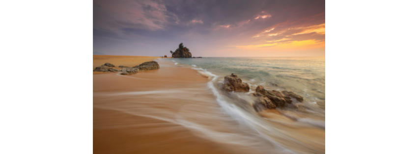 Waves washing over sand at sunset, with a rock stack offshore