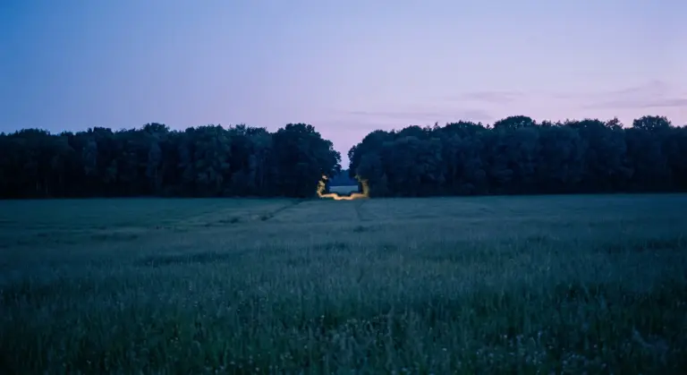 A dark blue field at dusk with a single lit doorway in the distant treeline